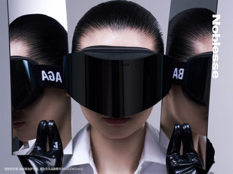 A woman poses in reflective goggles in a studio. Black gloves hold up mirrored panels with logo reflections. Fashion shot.