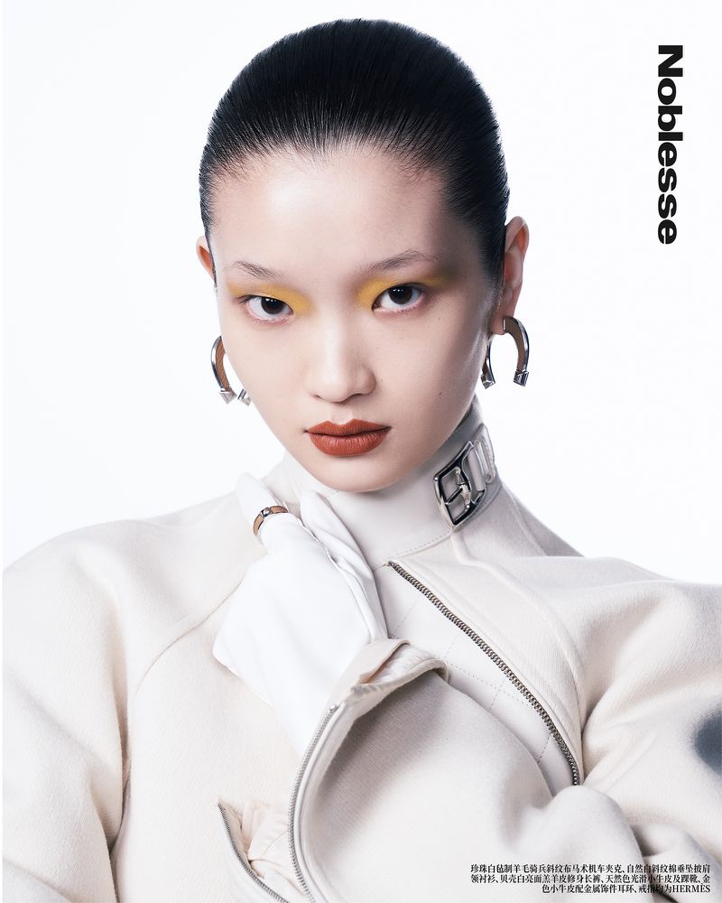 Close-up of an Asian woman with yellow eyeshadow and red lipstick wearing a white jacket and silver earrings in a studio shot.