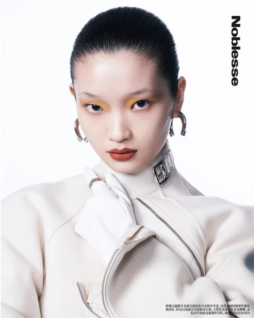 Close-up of an Asian woman with yellow eyeshadow and red lipstick wearing a white jacket and silver earrings in a studio shot.