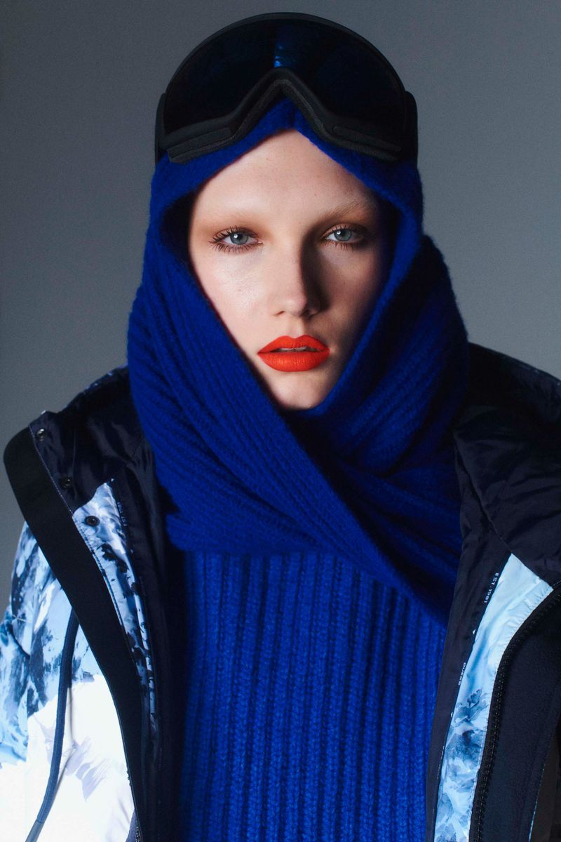 Close-up studio shot of a person in ski goggles, a bright blue scarf, and a jacket against a gray background for fashion.