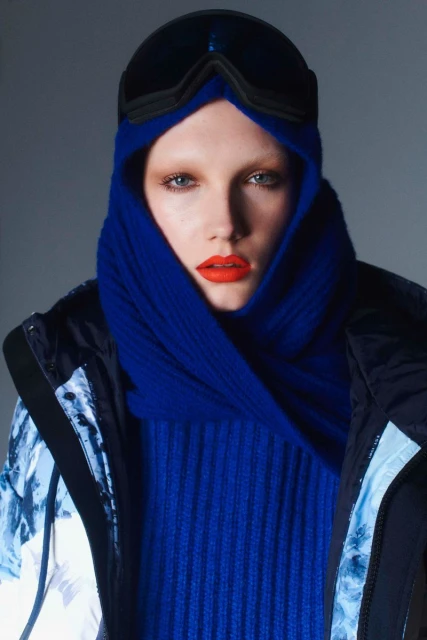 Close-up studio shot of a person in ski goggles, a bright blue scarf, and a jacket against a gray background for fashion.