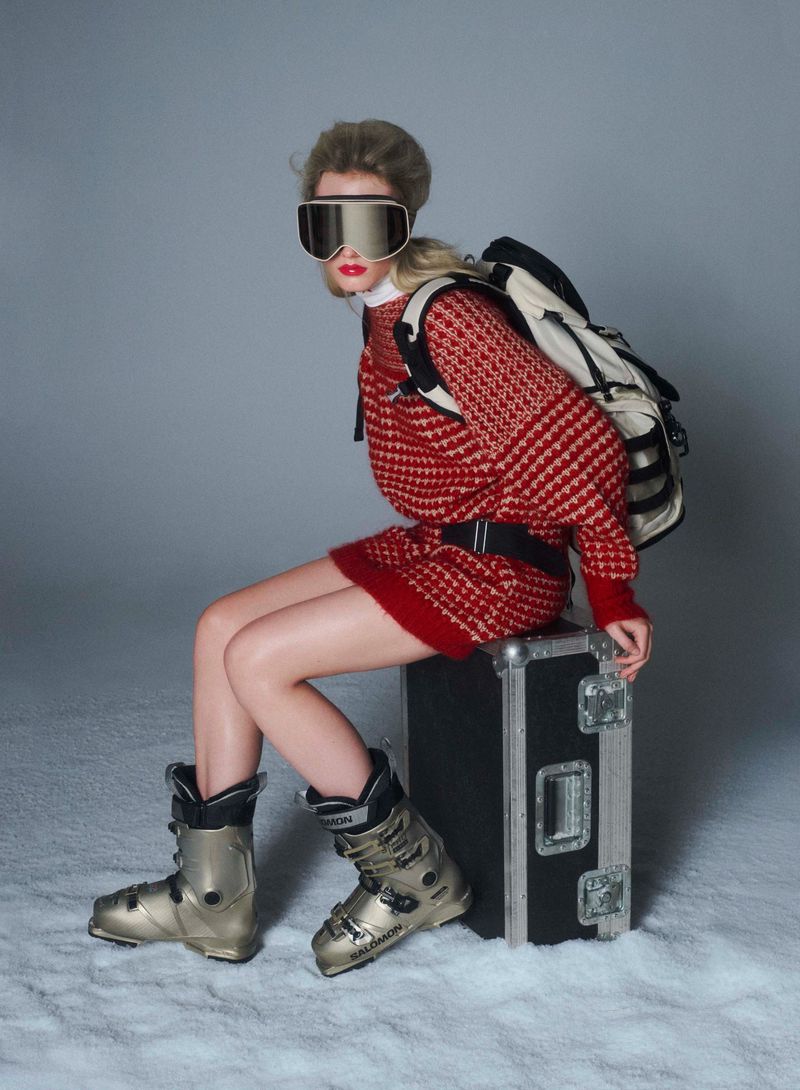 Blonde woman in red patterned sweater dress, ski goggles, boots, and backpack sits on black case in a snowy photo studio.