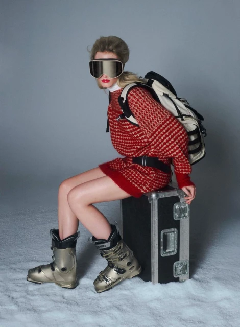 Blonde woman in red patterned sweater dress, ski goggles, boots, and backpack sits on black case in a snowy photo studio.