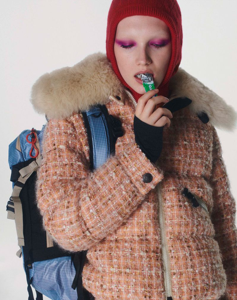 Woman in red balaclava and peach puffer jacket eating a snack, wearing backpack against a white backdrop, fashion editorial photography.