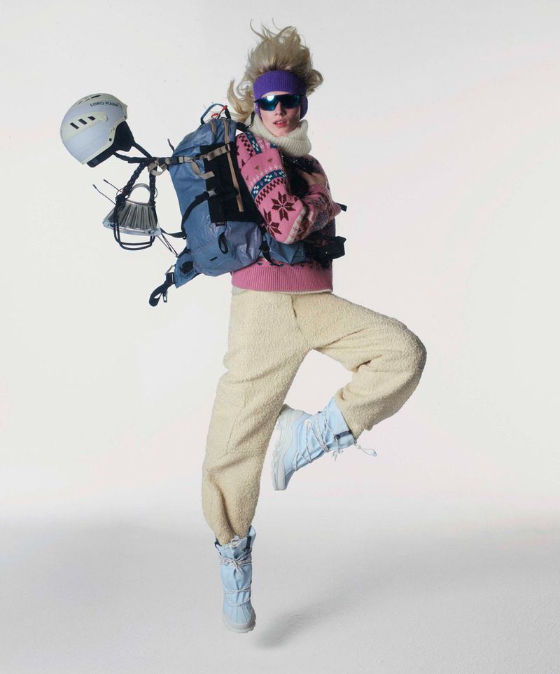 A person jumping in a white studio wearing a ski outfit, including a helmet, backpack, pink sweater, and boots, against a white background.