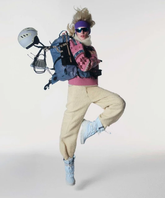 A person jumping in a white studio wearing a ski outfit, including a helmet, backpack, pink sweater, and boots, against a white background.