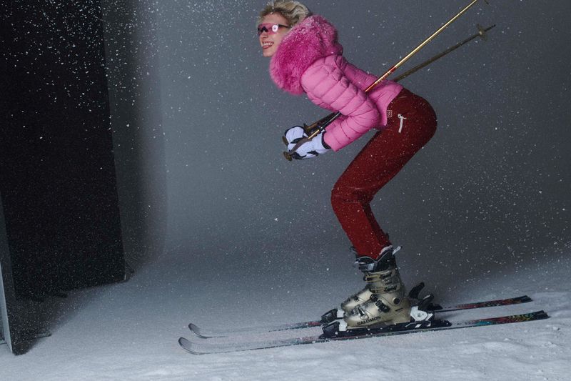 Stylish woman in pink jacket skiing downhill on snow with poles. Woman in ski apparel enjoying winter sport in an indoor snow setting.
