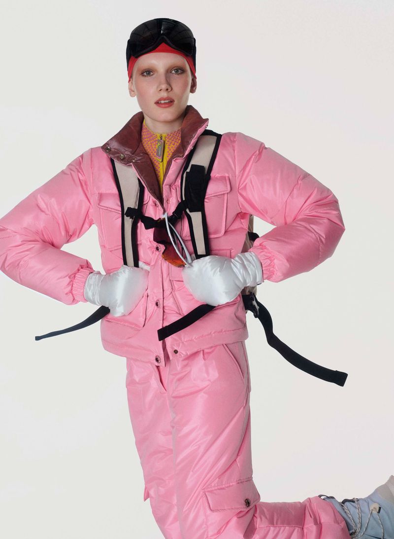 A model wearing a pink ski suit and black ski goggles poses in a studio environment against a white backdrop.