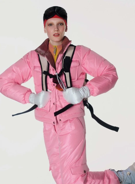 A model wearing a pink ski suit and black ski goggles poses in a studio environment against a white backdrop.