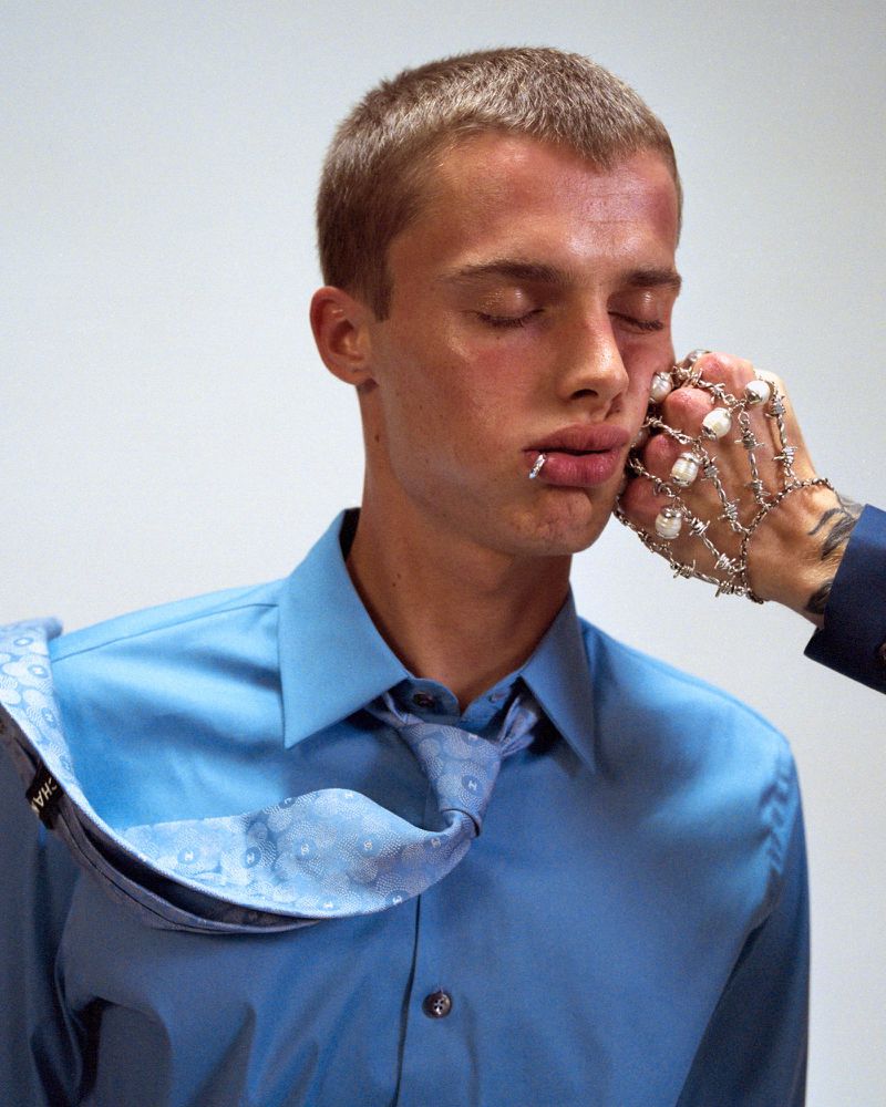 Young man with closed eyes, wearing a blue shirt and tie, is being punched in the face by a hand wearing wire jewelry.