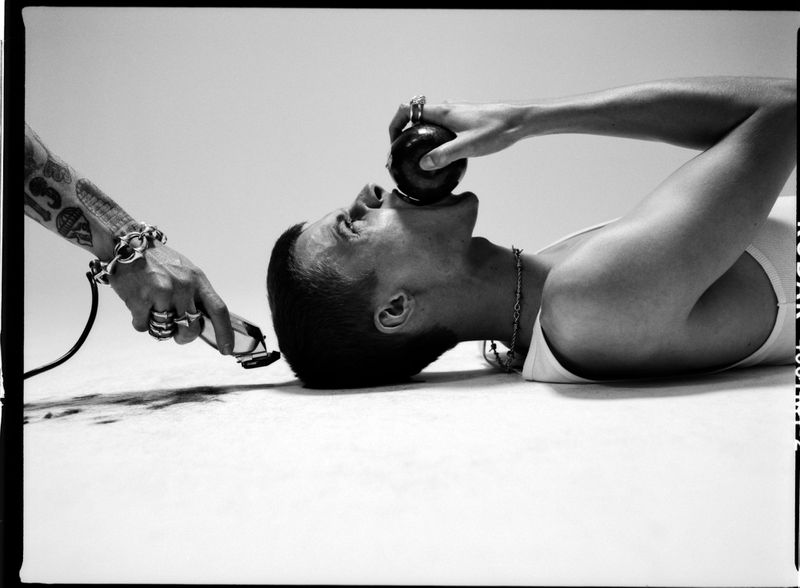 Black-and-white photo of a man lying down with an apple in his mouth, and an arm holding a pair of clippers next to his head.