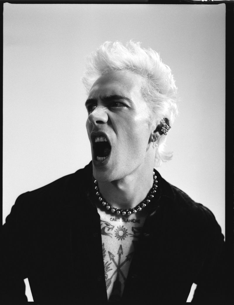 Monochrome close-up of a man with spiky white hair, a beaded necklace, and chest tattoos, shouting with an open mouth against a plain backdrop.