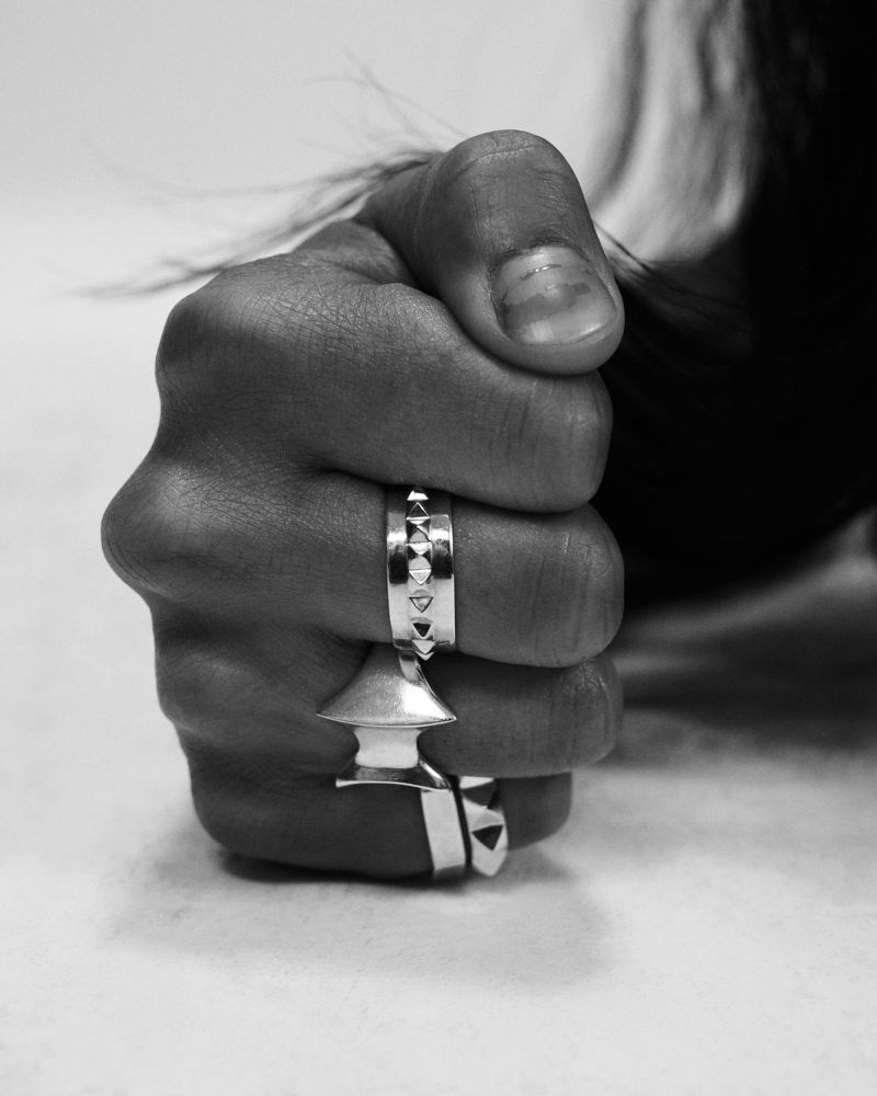 Close-up studio shot of hand wearing three silver rings, gripping dark hair against neutral background, minimal and fashionable.