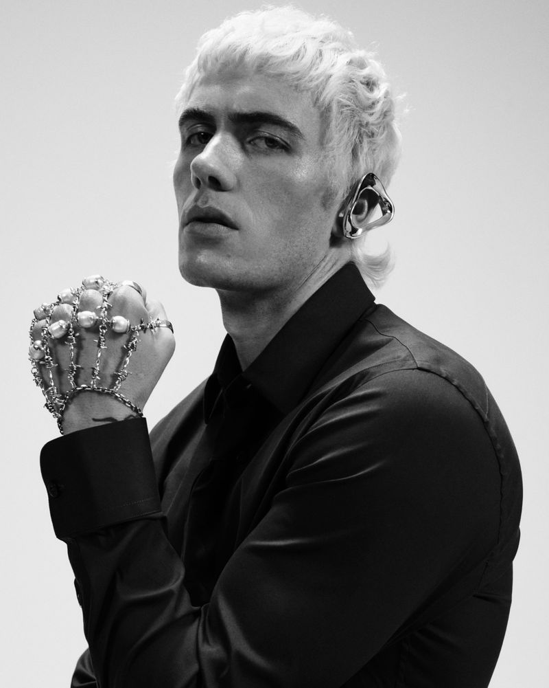 Monochrome portrait of a platinum-haired man wearing avant-garde jewelry, a black button-down shirt, and posing against a light backdrop.