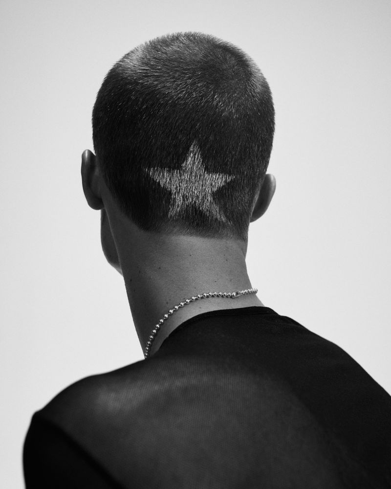 Monochrome studio shot of a person's head and shoulders with a star shaved into the back of their short hair, wearing a beaded necklace.