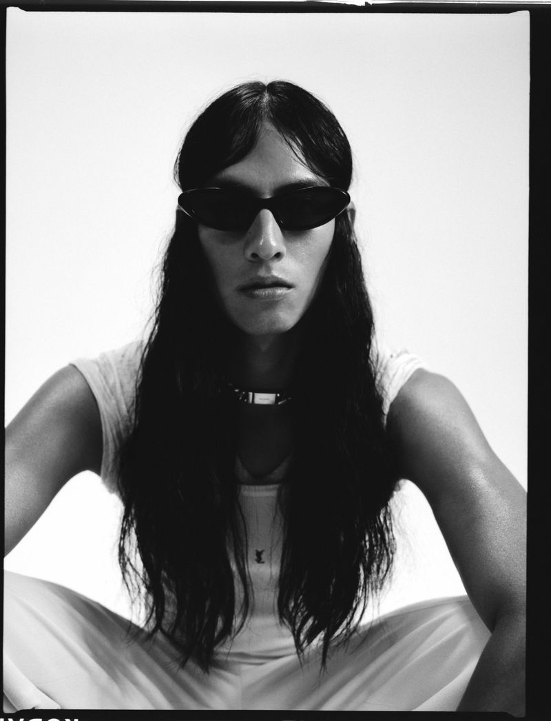 Person with long hair and sunglasses sits with legs apart against a white background in a studio portrait. A metal collar adorns the neck.