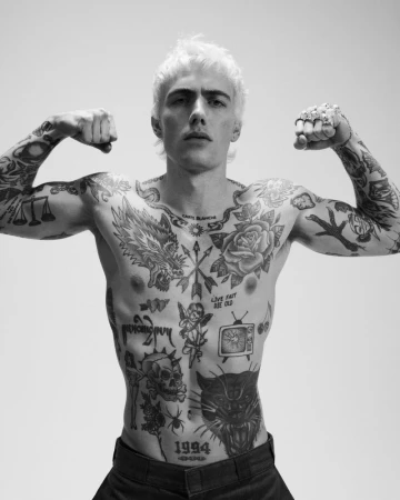 Black-and-white portrait of a young man with a bleached mullet, flexing arms and showcasing extensive tattoos, set against a bright background.