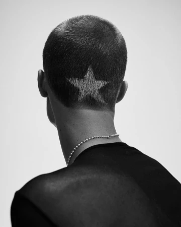 Back view of a man with a dark buzz cut featuring a bleached star design at the nape, wearing a black shirt and silver chain necklace.