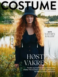 Model with long red hair wearing a dark jacket and cloche hat outdoors by a lake for Costume Norway magazine cover.