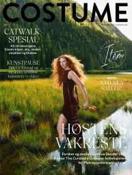 Model with long red hair wearing a dark sleeveless dress and tall boots walking through a grassy river landscape