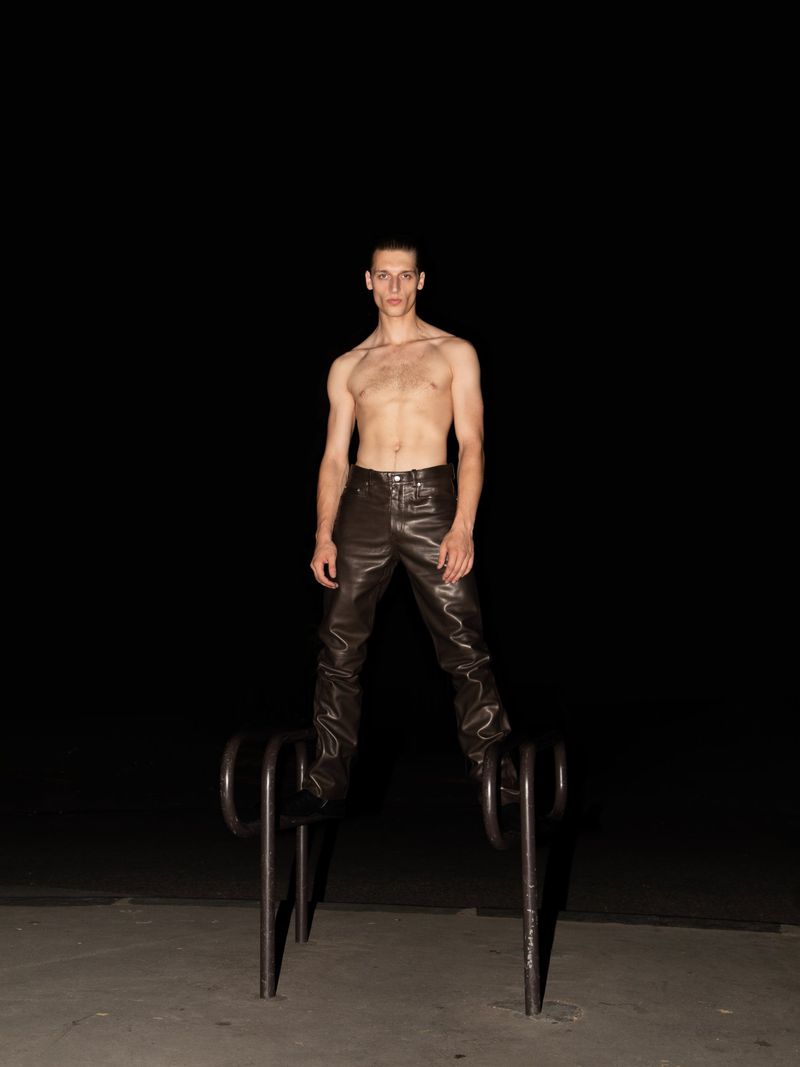 A slender shirtless man wearing leather pants stands atop a black metal bicycle rack outdoors against a stark black background in studio lighting.