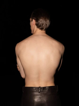 Shirtless man seen from behind wearing dark leather trousers against a plain black background.