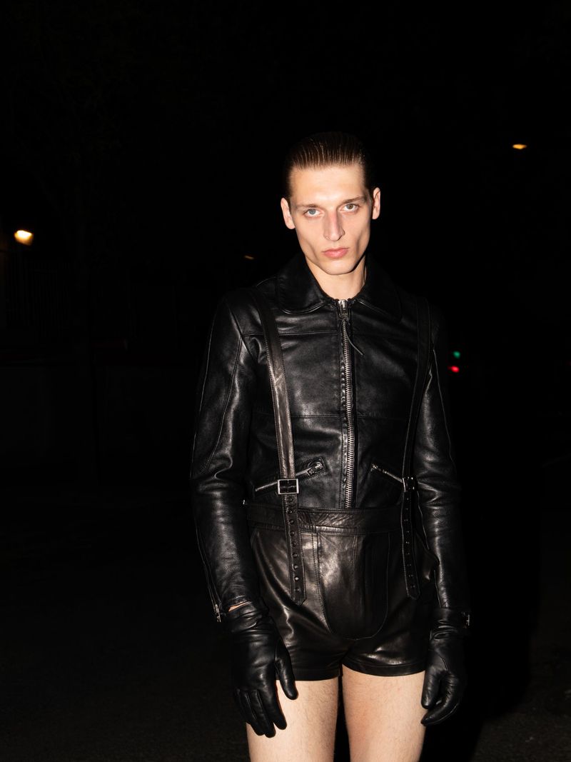 A male model poses in a black leather jacket and shorts, wearing leather gloves, against a dark, blurred background in an outdoor setting.
