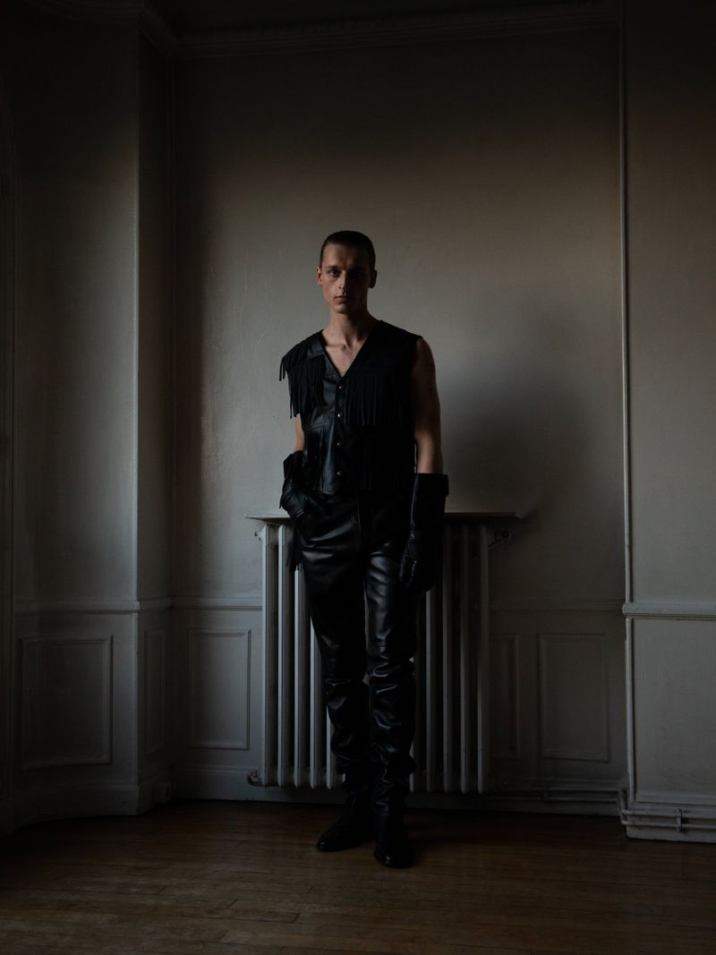 A slim adult male stands indoors against a plain wall, wearing a black leather fringed vest, black pants, boots, and gloves. 