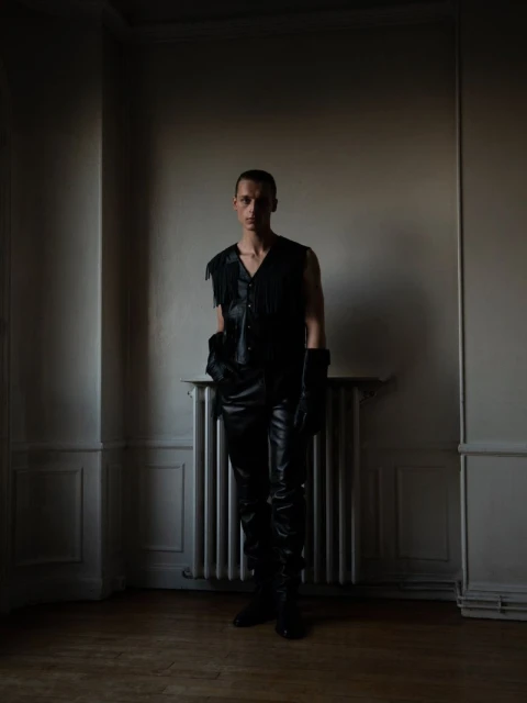 A slim adult male stands indoors against a plain wall, wearing a black leather fringed vest, black pants, boots, and gloves. 