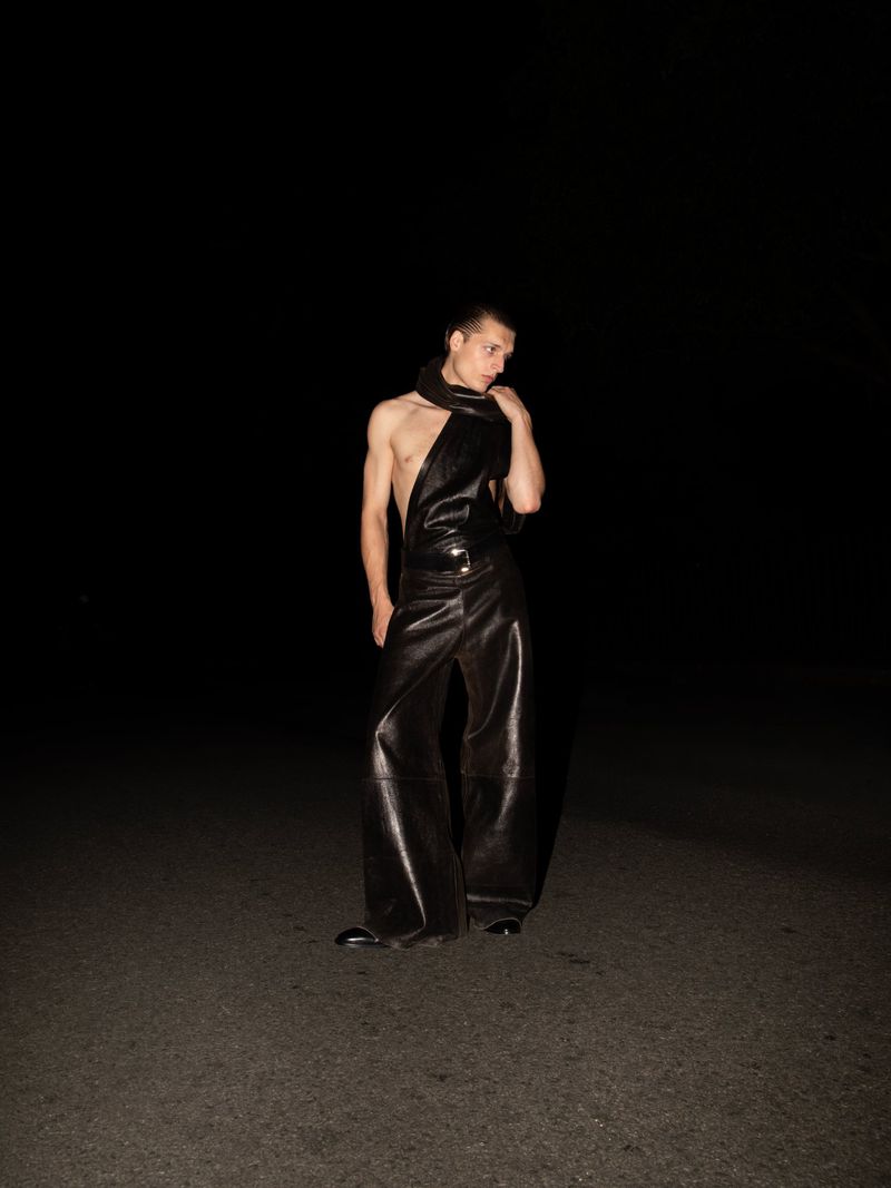 An adult male model poses in the darkness wearing a dark, one-piece garment with a scarf and black dress shoes on an asphalt surface.