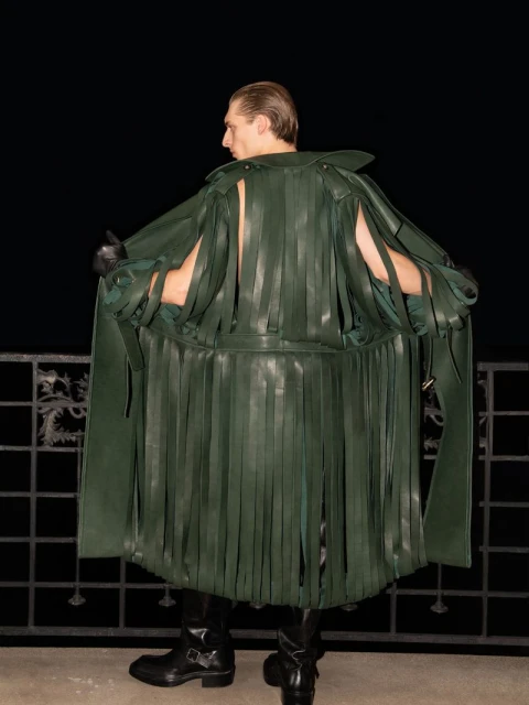 Back view of a slender male fashion model wearing a fringed green leather coat, black gloves, and black boots against a dark background.