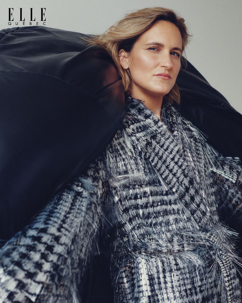 Fashion editorial portrait of a woman with blond hair wearing a textured black and white coat, against a neutral backdrop.
