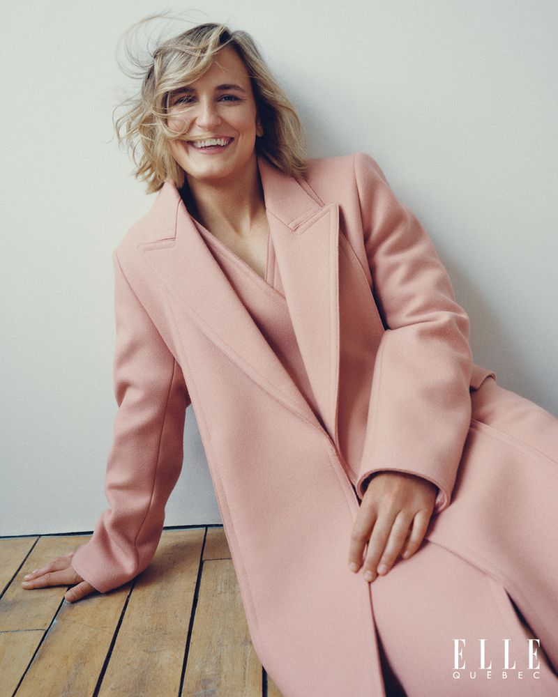 Smiling woman in a pink coat poses indoors against a white wall, radiating confidence and fashion-forward style in a portrait shot.
