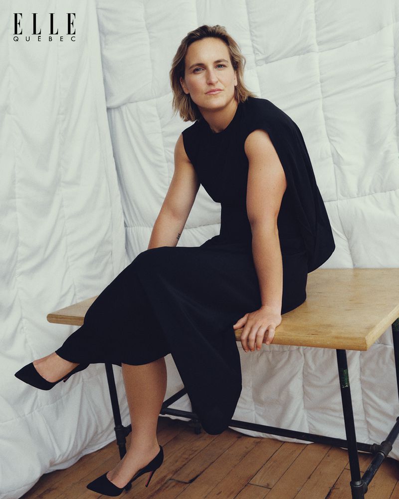 A blonde woman wearing a black dress sits on a wooden table with black heels in front of a white quilted wall, posing for a fashion editorial.