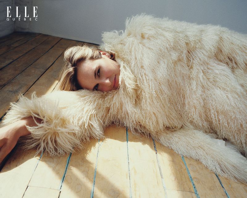 A woman is laying on a wooden floor wearing a large, white, fuzzy jacket. The Elle Quebec magazine logo is visible in the top-left.