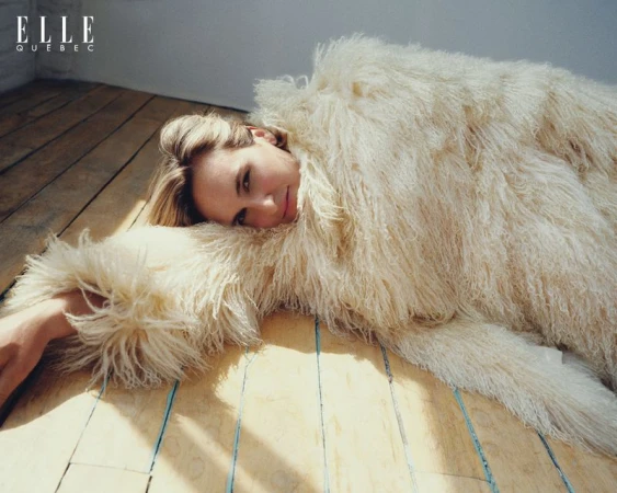 A woman is laying on a wooden floor wearing a large, white, fuzzy jacket. The Elle Quebec magazine logo is visible in the top-left.