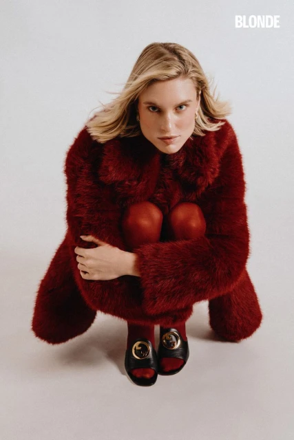 Blonde-haired woman in a red fur coat poses in a photo studio wearing red tights and black sandals, captured in a high-angle shot.