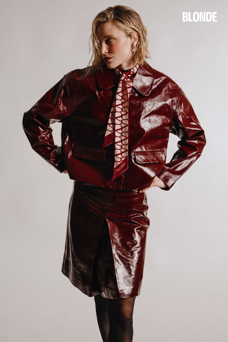 Fashion shot of a blonde woman in a shiny red jacket and skirt, posed against a neutral background, wearing a patterned scarf tie.