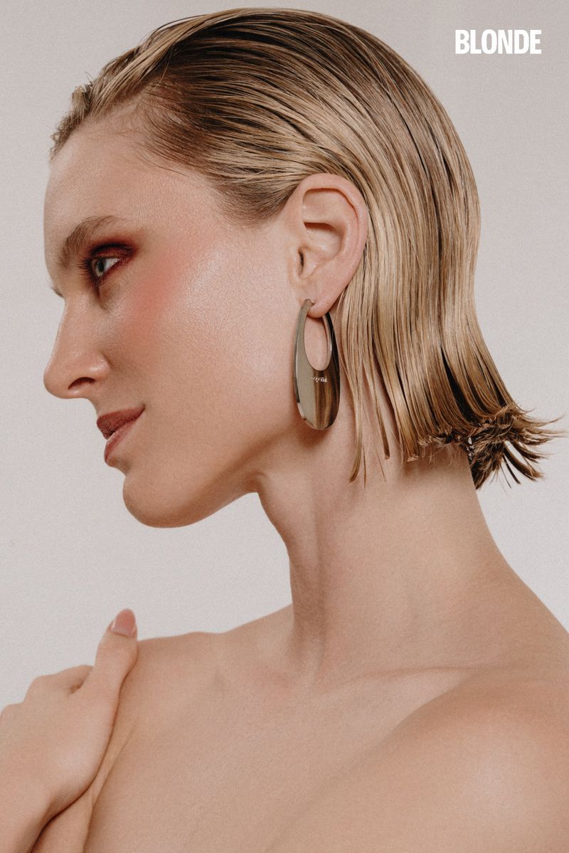 Blonde-haired woman with shoulder-length wet-look hair poses against a neutral background with bold makeup and large silver earrings.