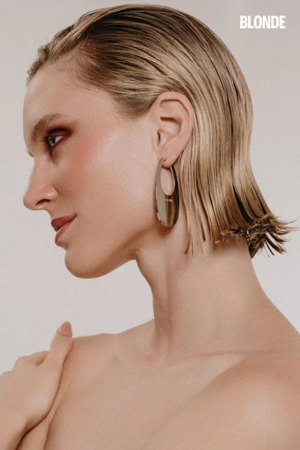 Blonde-haired woman with shoulder-length wet-look hair poses against a neutral background with bold makeup and large silver earrings.