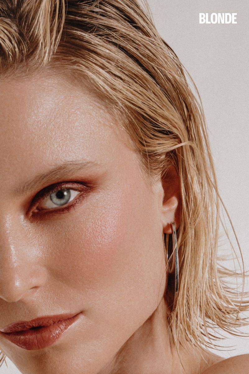 Close-up portrait of a blonde woman with light skin, blue eyes, and neutral makeup, wearing silver hoop earrings against a white background.