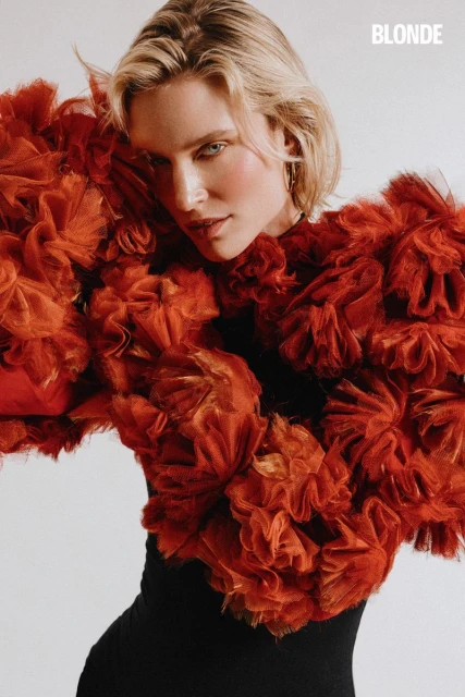 Portrait of a blonde adult female wearing a black top and a voluminous, frilly orange jacket in front of a white background.