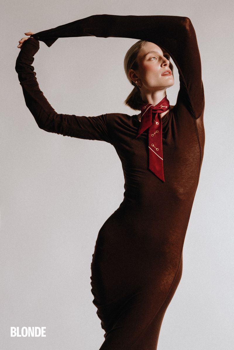 Fashion shot of a woman in a brown dress with a red neck scarf, arms raised, on a white background. Studio lighting. Editorial style.