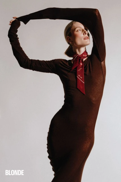 Fashion shot of a woman in a brown dress with a red neck scarf, arms raised, on a white background. Studio lighting. Editorial style.