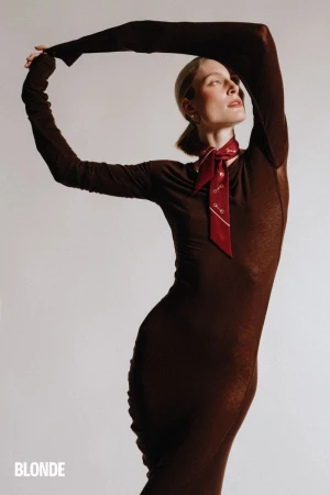 Model wearing a long-sleeved brown dress and red neck scarf posing against a plain neutral background with arms raised overhead