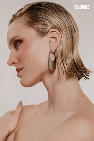 Profile portrait of a woman with wet slicked hair wearing a large metallic hoop earring and red eye makeup against a white backdrop.