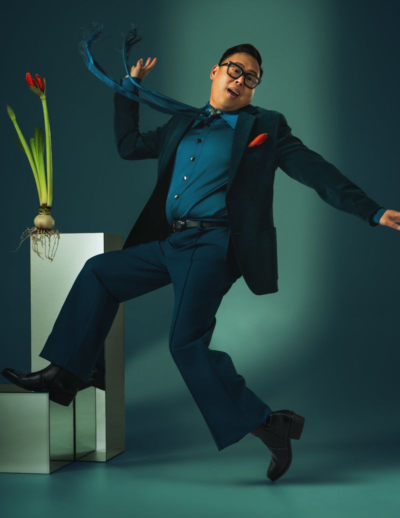 Man in a teal suit strikes a dynamic pose next to tulips on a white block, against a teal backdrop in a studio, creating a vibrant and lively image.