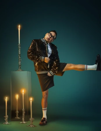 Man in sequined jacket with leg raised, standing near candles on a green backdrop, wearing a collared shirt, loafers, and socks.