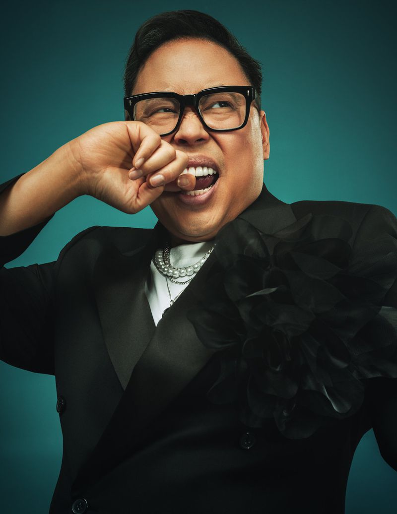 Portrait of adult male with glasses and a black suit. He is holding a lozenge in his mouth against a teal studio background.