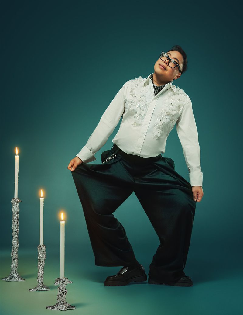 Well-dressed person in wide-legged pants poses with lit candles against a blue-green backdrop in a full studio shot, looking upward.